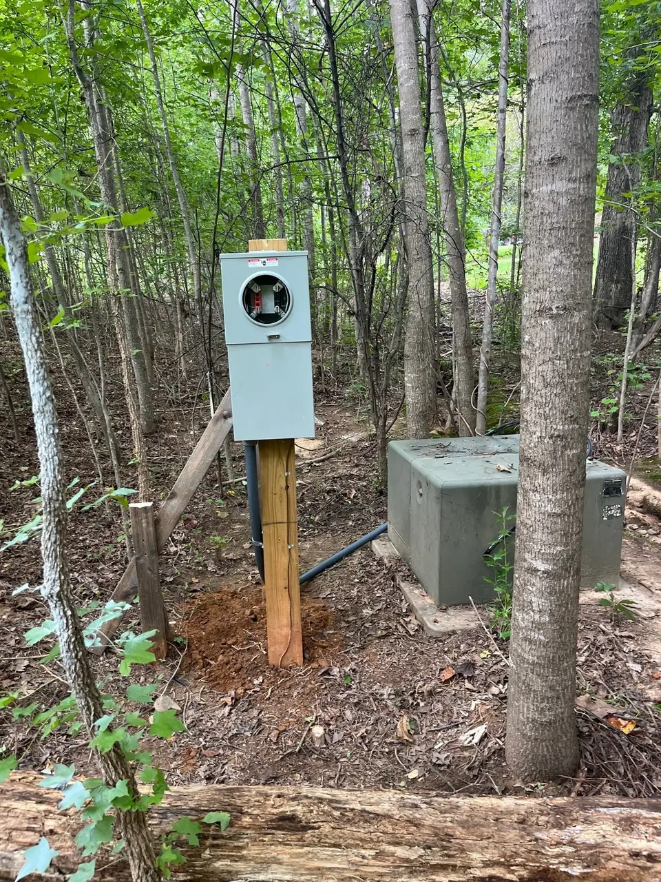 A new meter base mounted on a wooden post beside a utility transformer box in a wooded lot.